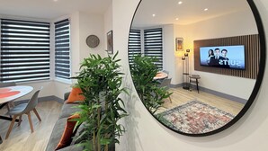 A modern living room with a round mirror reflecting the space, a dining area with a table and chairs, and a TV mounted on the wall displaying a show. - Humberside Apartments (Cleethorpes)