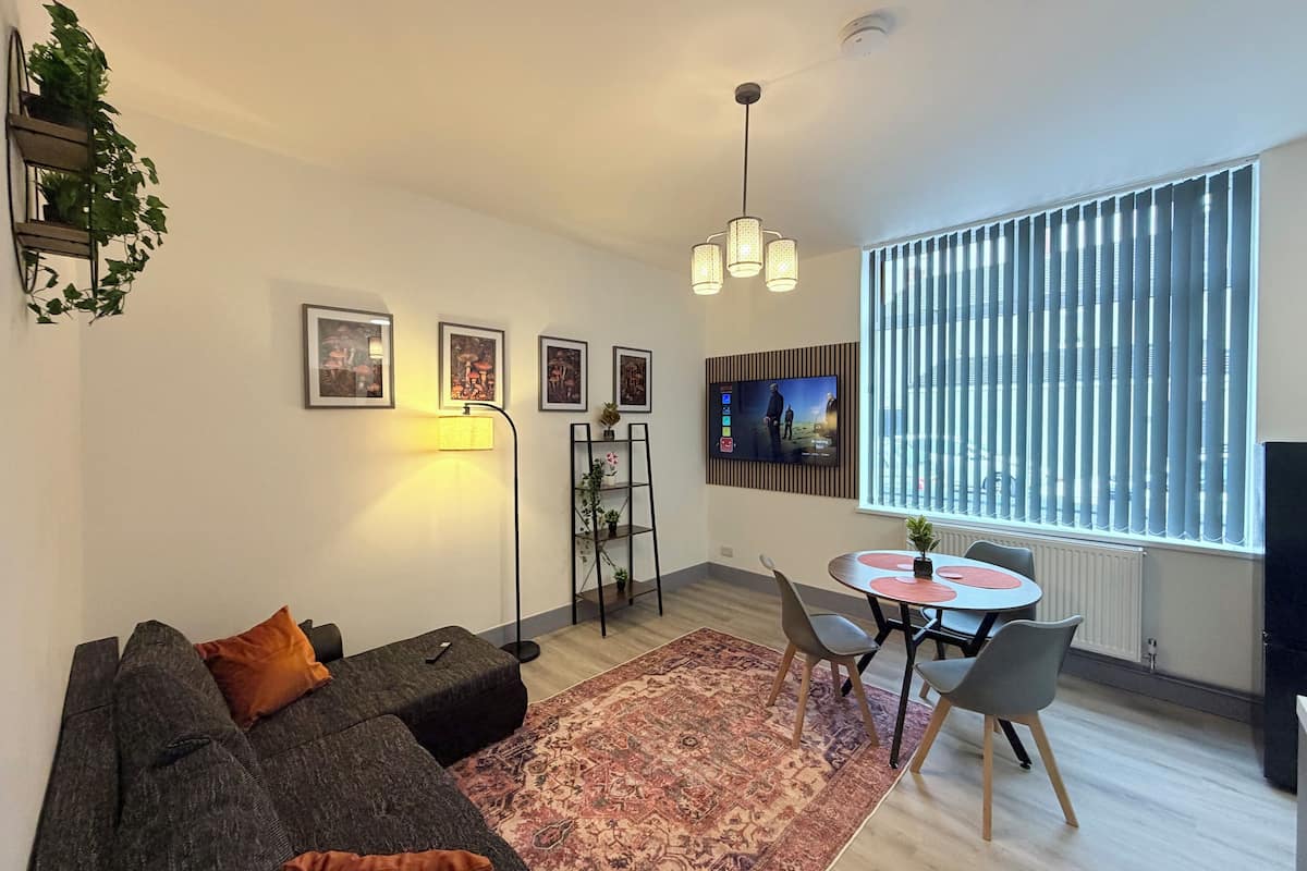 A modern living room with a grey sectional sofa, a patterned rug, a dining area with a small table and chairs, a TV mounted on the wall, and a large window with blinds.