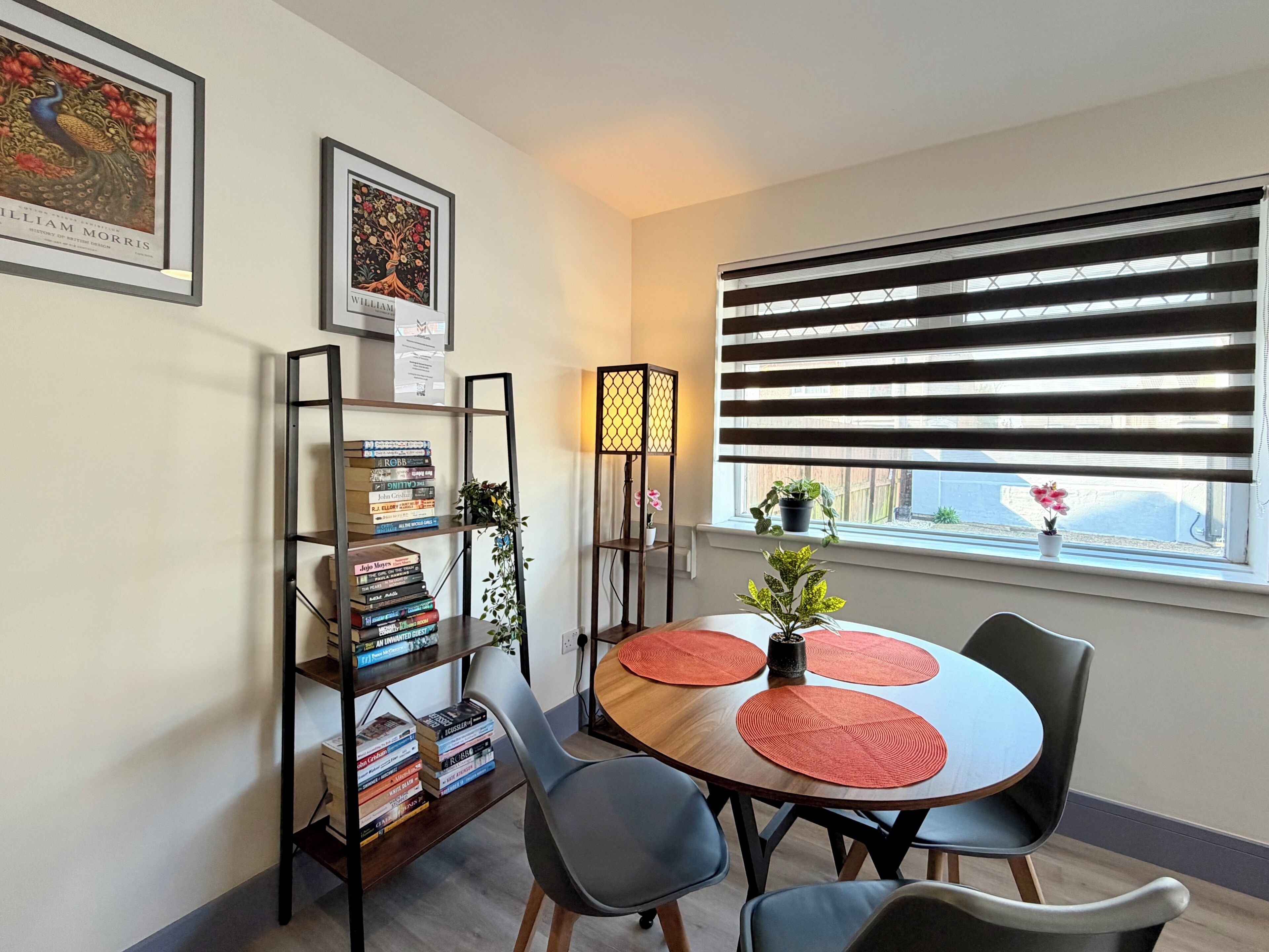 A dining area with a round table, chairs, a bookshelf, and a window with blinds.