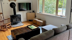 A living room with a black wood-burning stove, a flat-screen TV mounted on the wall, a grey sofa, and a wooden coffee table. - Charming Berkshire Bungalow: Deck, Firepit, Stargazing (Egremont)