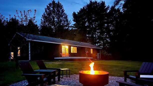 A cozy outdoor seating area with a fire pit, surrounded by chairs and a table, in front of a house with a lit interior. - Charming Berkshire Bungalow: Deck, Firepit, Stargazing (Egremont)