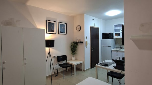 Una habitación de hotel con cama, escritorio, silla, armario, una pequeña mesa con una planta, un reloj y una kitchenette.