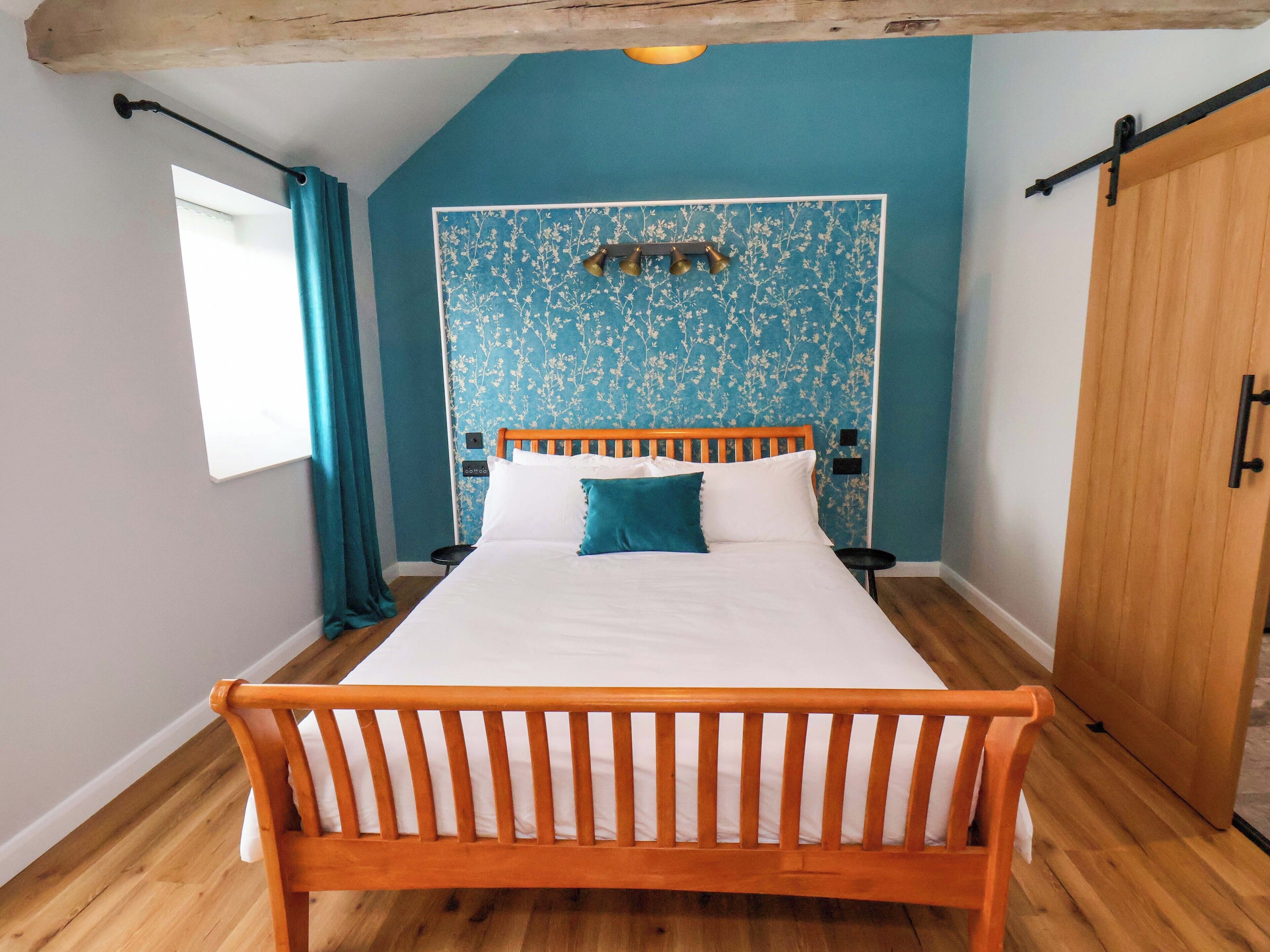 Une chambre à coucher avec un lit en bois, un papier peint à motifs floraux bleus, une armoire en bois et une petite fenêtre avec des rideaux.