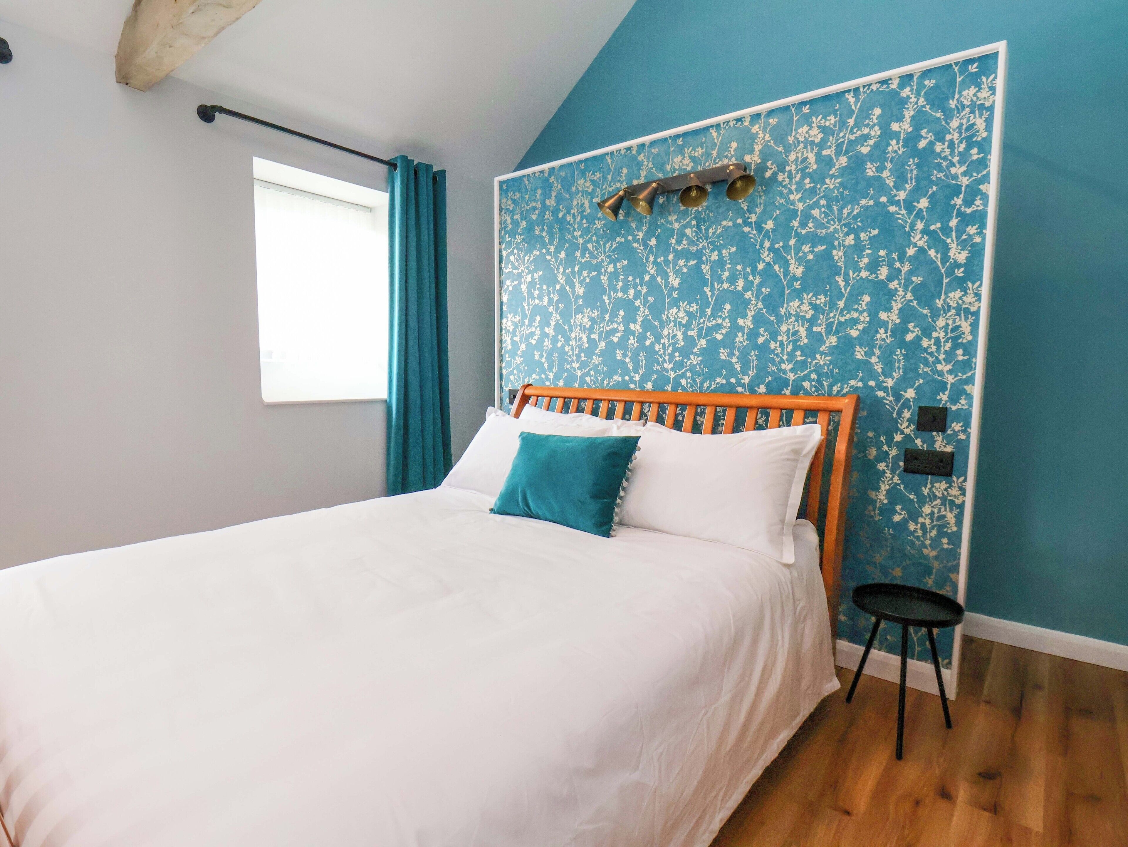 Een slaapkamer met een houten bed, witte beddengoed, een hoofdeinde met een blauw bloemenmotief, een klein zwart krukje en een raam met gordijnen.