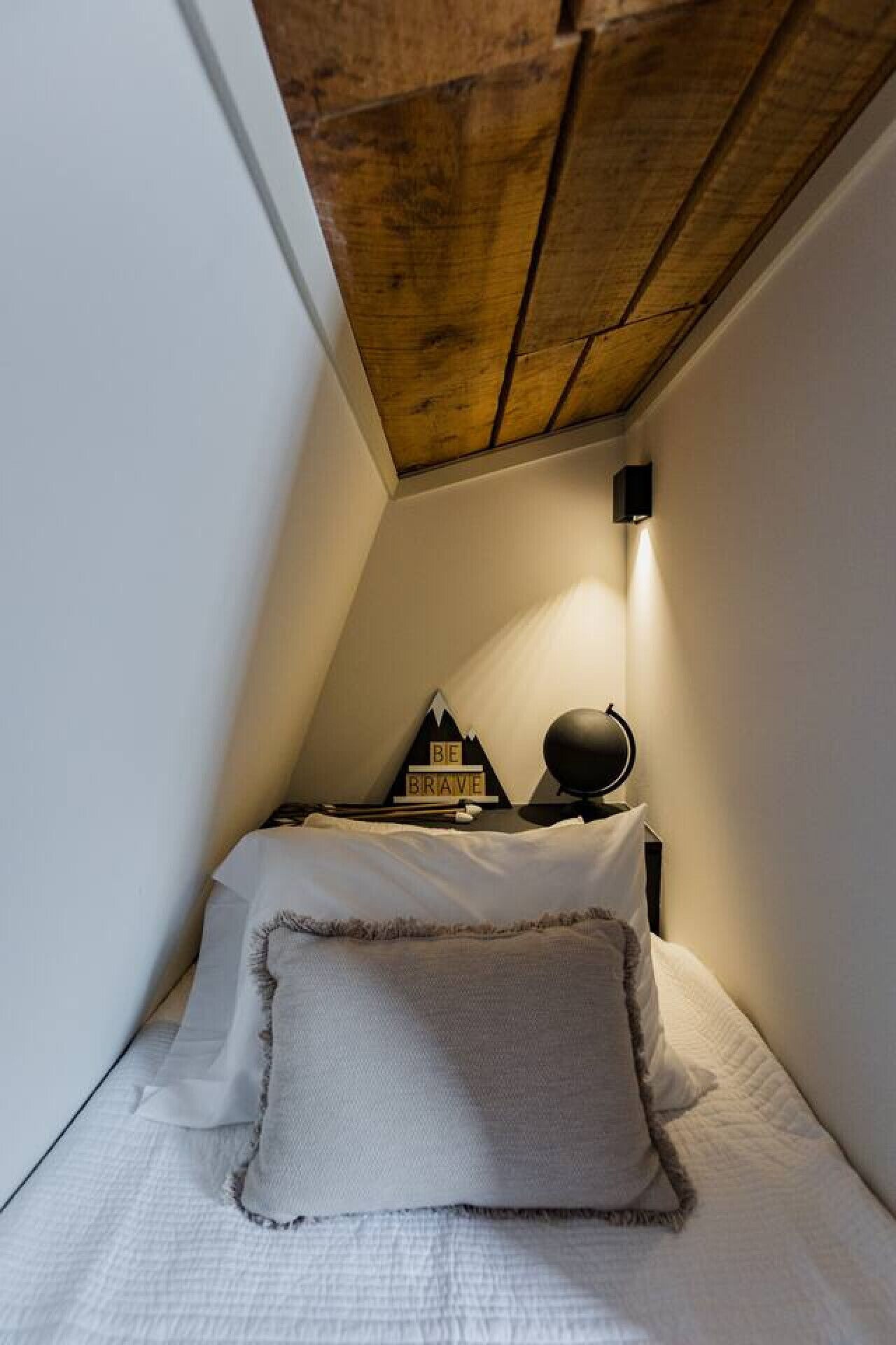 Ein kleines, gemütliches Schlafzimmer mit schrägen Wänden, einem Bett mit weißer Bettwäsche, einer Nachttischlampe und einem motivierenden Spruch an der Wand.