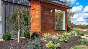 Una casa moderna de madera con un ventanal grande, rodeada de un jardín ajardinado con diversas plantas y arbustos.