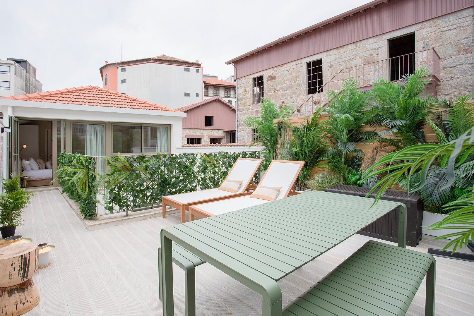 A rooftop terrace with a green table and two lounge chairs, surrounded by lush greenery and a stone building in the background.