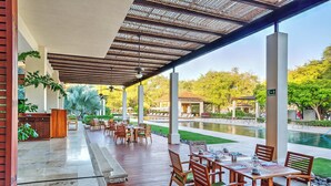 An outdoor patio with wooden tables and chairs, a pool, and a covered area with a view of the pool and greenery. - House Reserva Conchal Llama del Bosque (Cabo Velas)