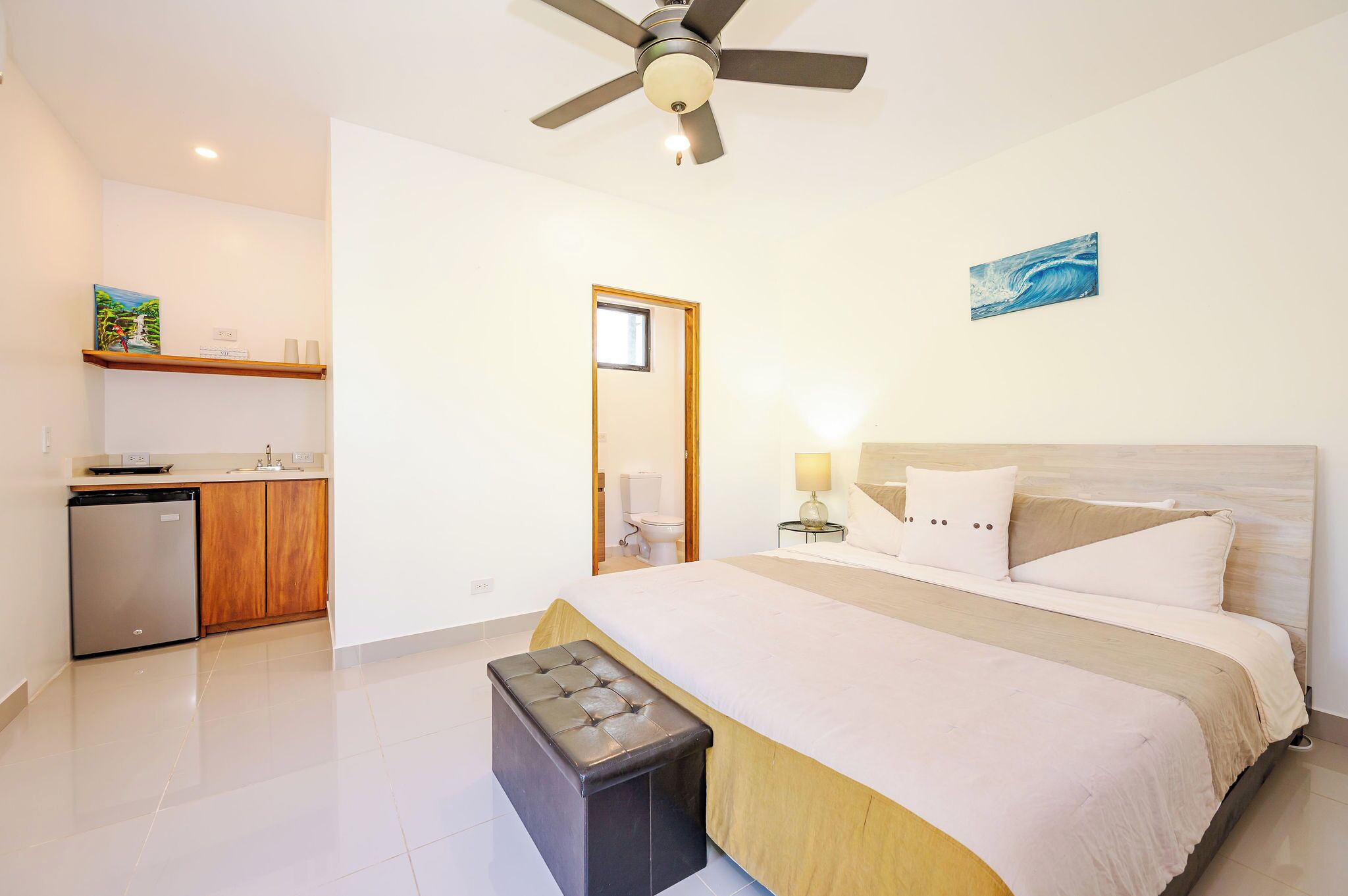 A bedroom with a bed, a bench, a ceiling fan, a nightstand, and a bathroom visible through an open door.
