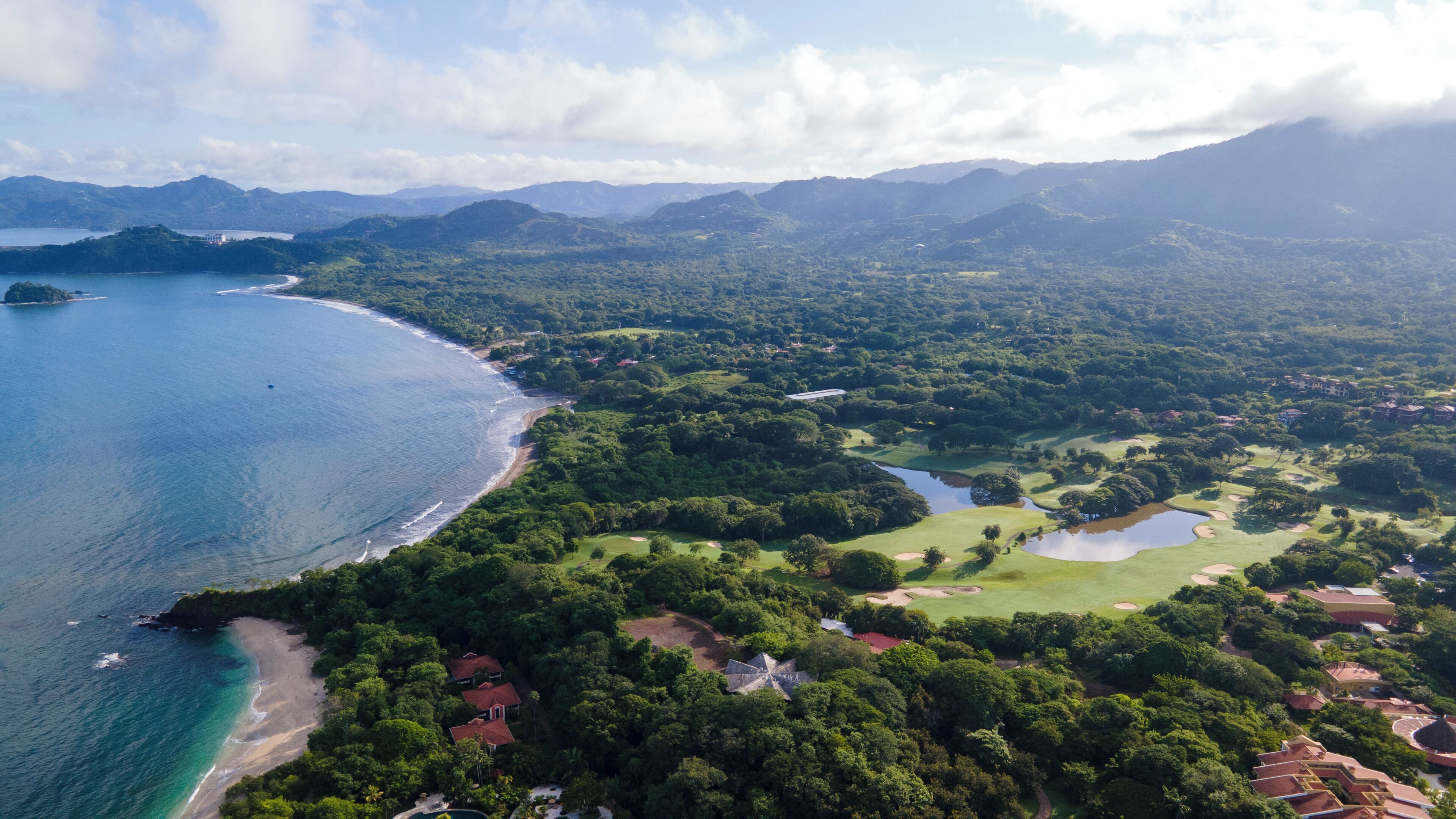 Vista aérea de una zona costera con playa, campo de golf y colinas boscosas circundantes.