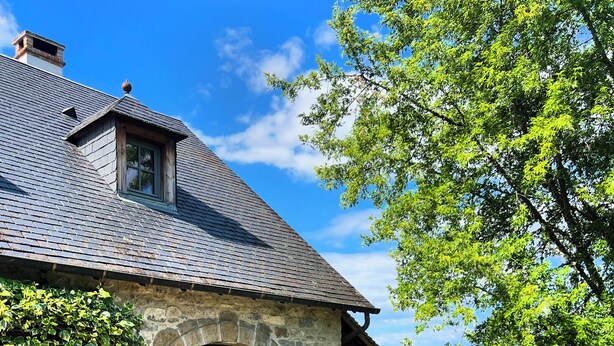 A stone building with a sloped roof, a covered patio with wooden furniture, and a view of the surrounding landscape. - La Blanchie Haute, Gîte de charme d'exception pour 2 en Vallée de la Dordogne (Martel)