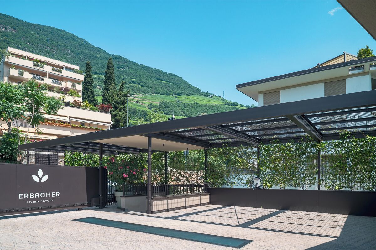 Un espace extérieur moderne doté d’un coin salon abrité, d’une enseigne indiquant « ERBACHER LIVING NATURE » et offrant une vue sur une colline.