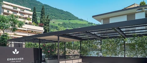 Un espace extérieur moderne doté d’un coin salon abrité, d’une enseigne indiquant « ERBACHER LIVING NATURE » et offrant une vue sur une colline.