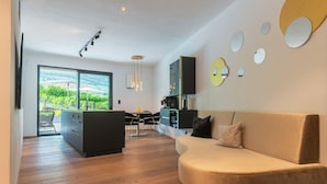 A modern living room with a curved sofa, a dining area with a table and chairs, and a kitchen area with a coffee machine. - Holiday Apartment ‘Erbacher – Gabi’s Suite’ with Mountain View, Sauna and Pool (Bozen)