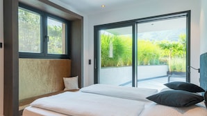 A bedroom with a large bed, a view of greenery through a sliding door, and a window with a view of trees. - Holiday Apartment ‘Erbacher – Gabi’s Suite’ with Mountain View, Sauna and Pool (Bozen)