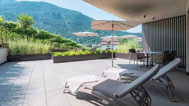 A terrace with lounge chairs, a table, and umbrellas, overlooking a mountainous landscape. - Holiday Apartment ‘Erbacher – Gabi’s Suite’ with Mountain View, Sauna and Pool (Bozen)