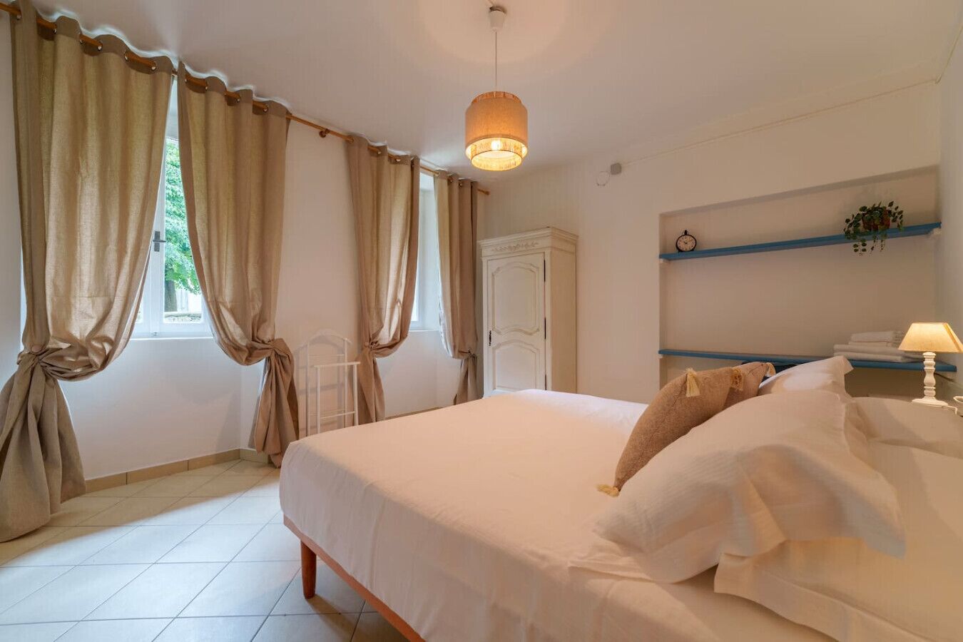 A bedroom with a bed, curtains, a bedside table with a lamp, a shelf with a clock, and a door.