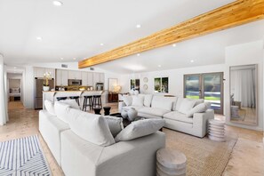 A modern living room with a large sectional sofa, a kitchen in the background, and a wooden ceiling beam. - The Dean - Modern Zen Retreat in Deepwell (Palm Springs)
