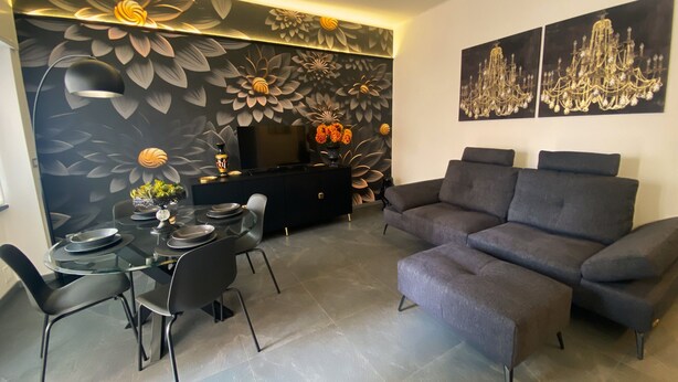 En moderne stue med en grå sofa, et spisebord med stole, et sort tv-møbel og et blomstret tapet.
