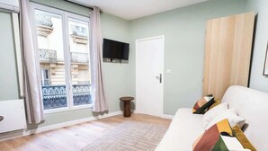 A room with a sofa, a small round table, a TV mounted on the wall, and a view of a building through a window with a curtain. - Gare de Lyon - charming modern studio (Paris)