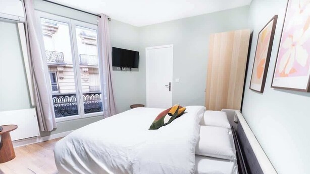 A bedroom with a bed, a television on the wall, and a window with a view of buildings. - Gare de Lyon - charming modern studio (Paris)