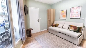 A living room with a sofa, a TV mounted on the wall, a door, and a window with a view of buildings. - Gare de Lyon - charming modern studio (Paris)