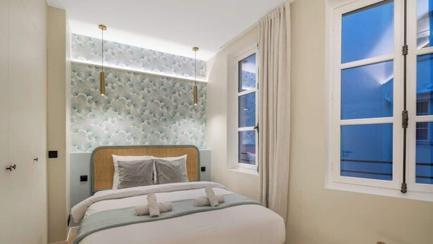 A bedroom with a bed, a window with curtains, a wall with a floral pattern, and a wooden bench. - Élégant 3 pièces climatisé au cœur du Marais (Paris)