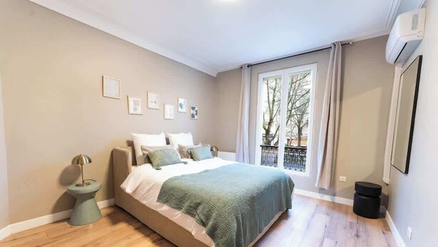Een slaapkamer met een bed, een nachtkastje, een raam met gordijnen en een luchtverkoeler aan de muur.