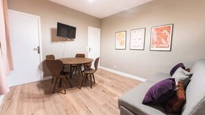 Een woonkamer met een grijze bank, een houten eettafel met stoelen en een flatscreen televisie aan de muur.