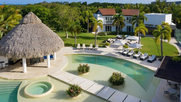 Un resort con piscina, sillas de descanso, una estructura de techo de paja y una casa grande con techo rojo.
