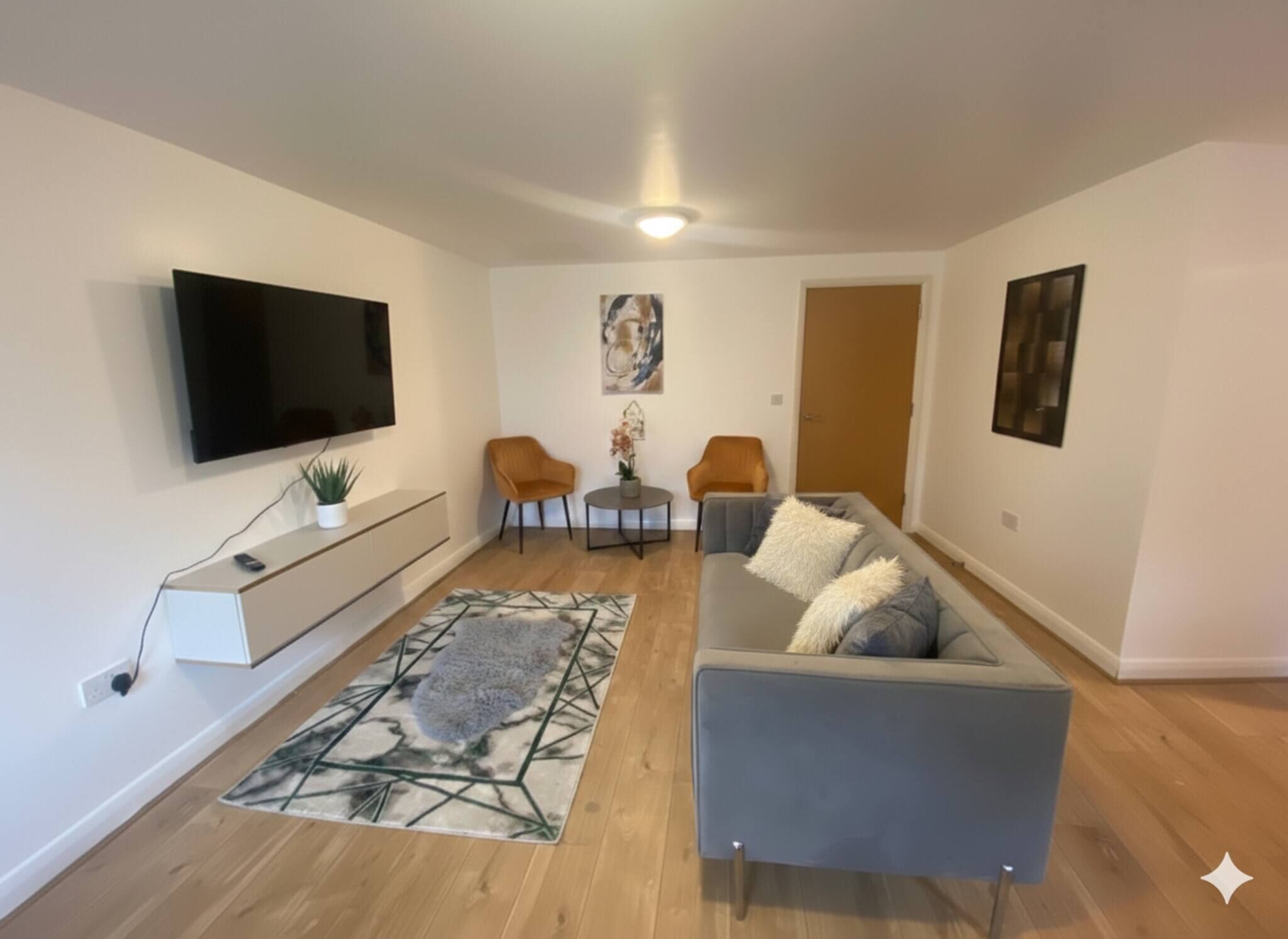 Un salon moderne avec un téléviseur à écran plat, un canapé gris, deux fauteuils, une table basse et un tapis à motifs.