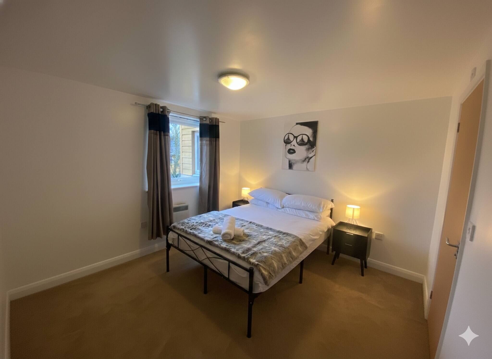 A bedroom with a bed, bedside lamps, a window with curtains, a radiator, and a wall-mounted artwork.