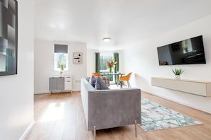 A modern living room with a grey sofa, a flat-screen TV, a dining area with a table and chairs, and a window with curtains. - Stylish 2 - Bed Manning Gardens (Croydon)
