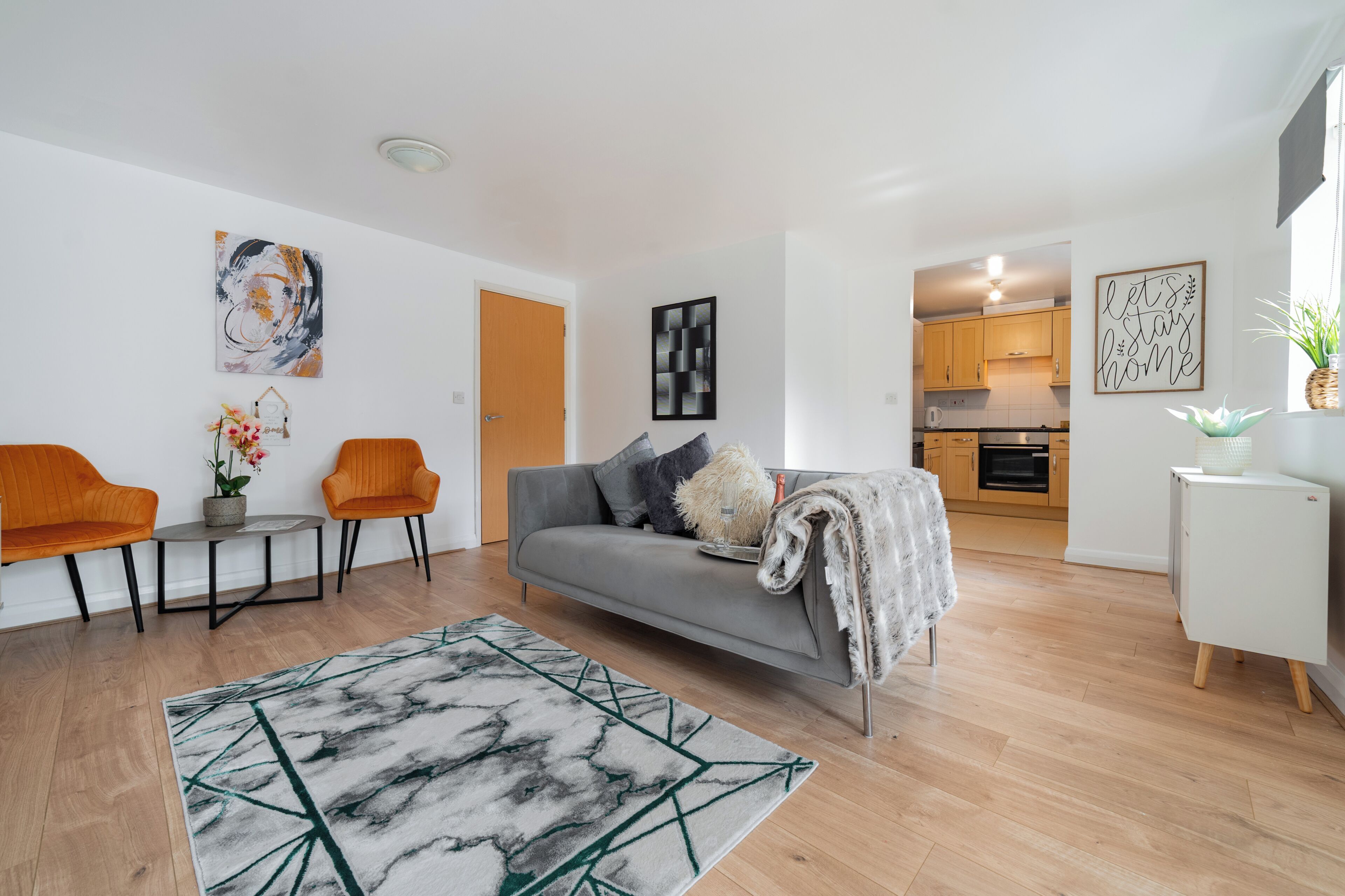 A modern living room with a grey sofa, orange chairs, a marble-patterned rug, and a kitchen in the background.