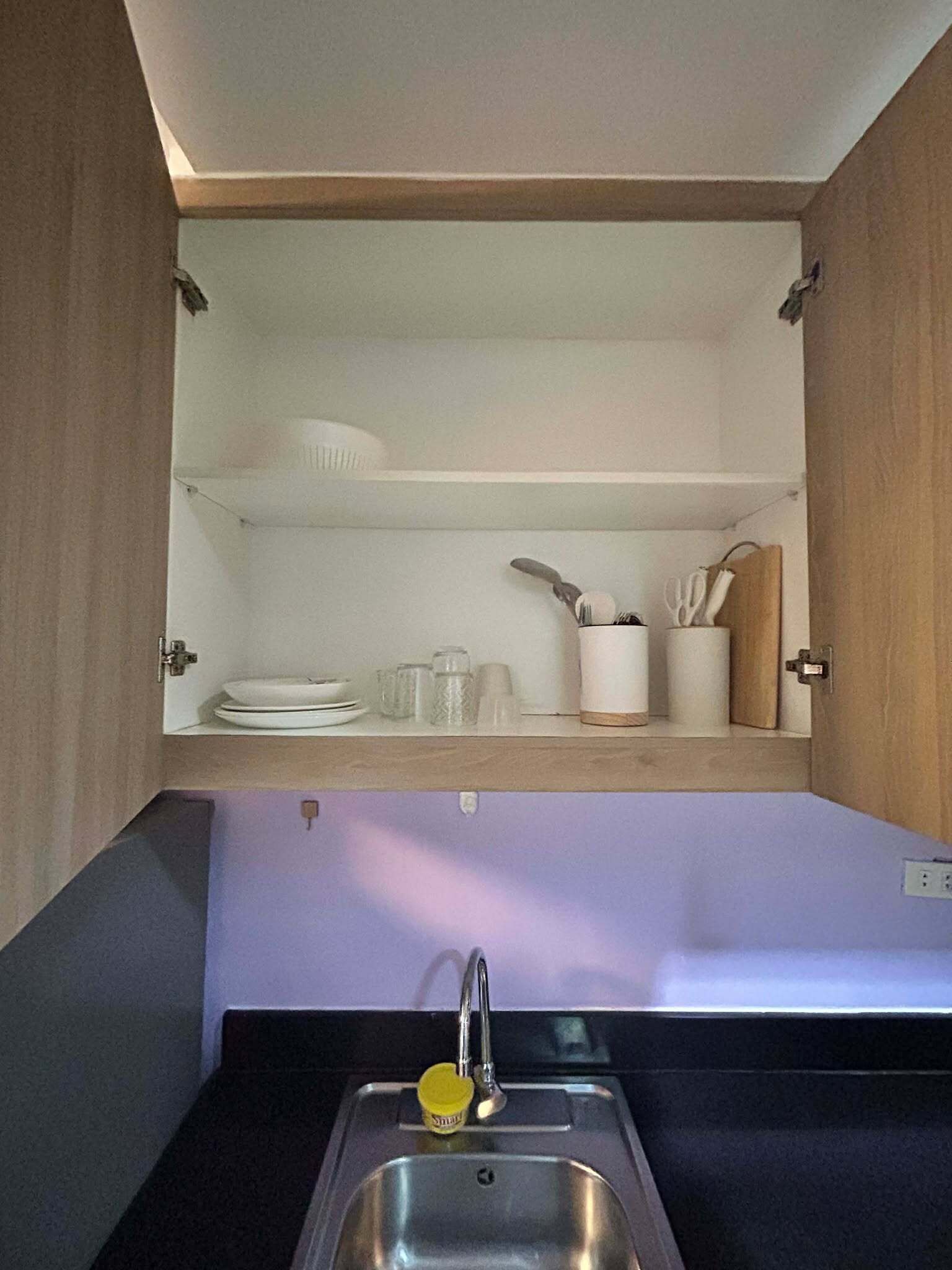 Una cocina con un lavamanos integrado, un estante sobre el lavamanos que contiene platos y utensilios, y una puerta de armario abierta que muestra el interior.