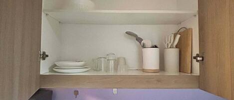 Una cocina con un lavamanos integrado, un estante sobre el lavamanos que contiene platos y utensilios, y una puerta de armario abierta que muestra el interior.