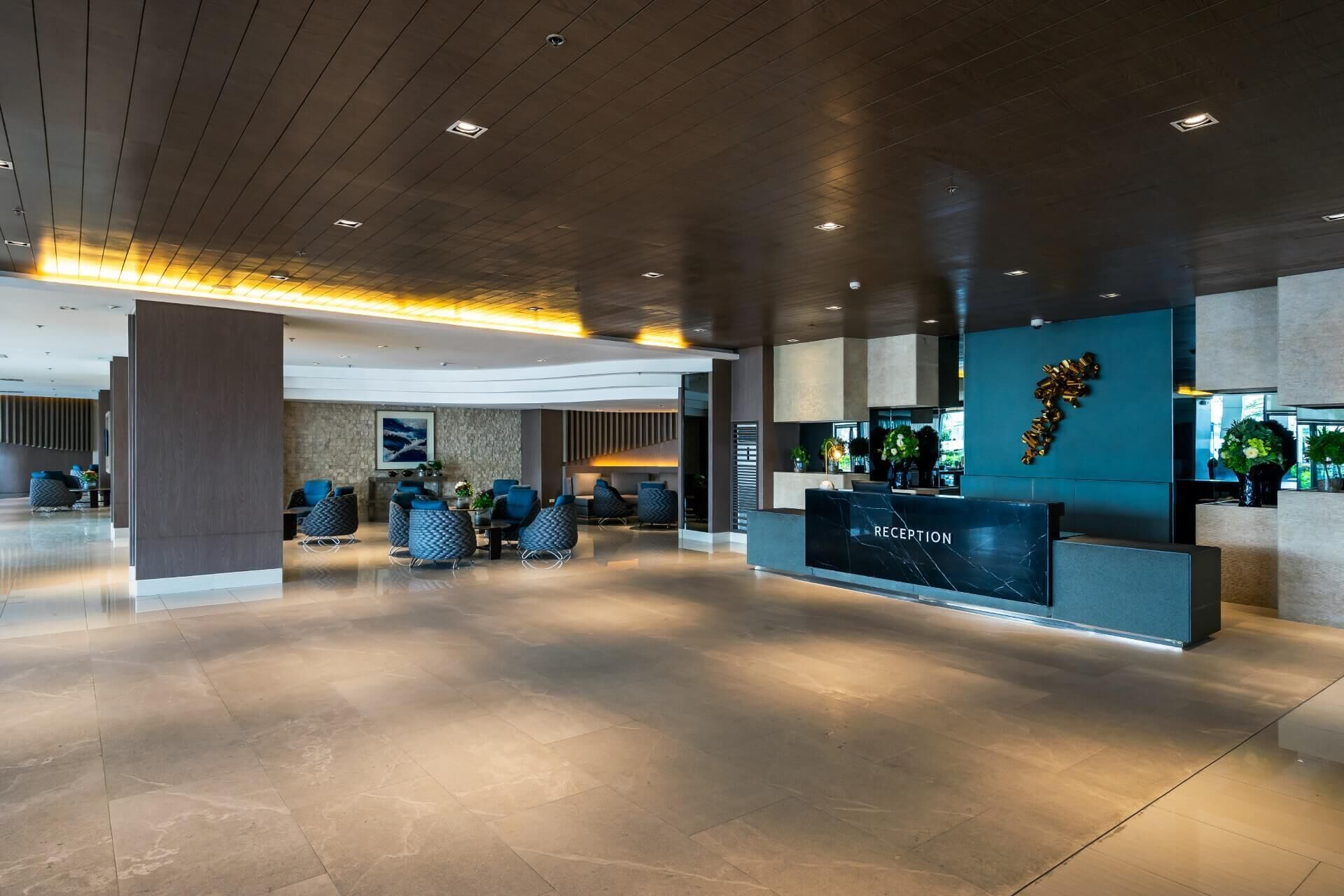 Un espace d’accueil moderne avec un comptoir de réception en marbre, un coin salon avec des fauteuils bleus et un élément décoratif mural.