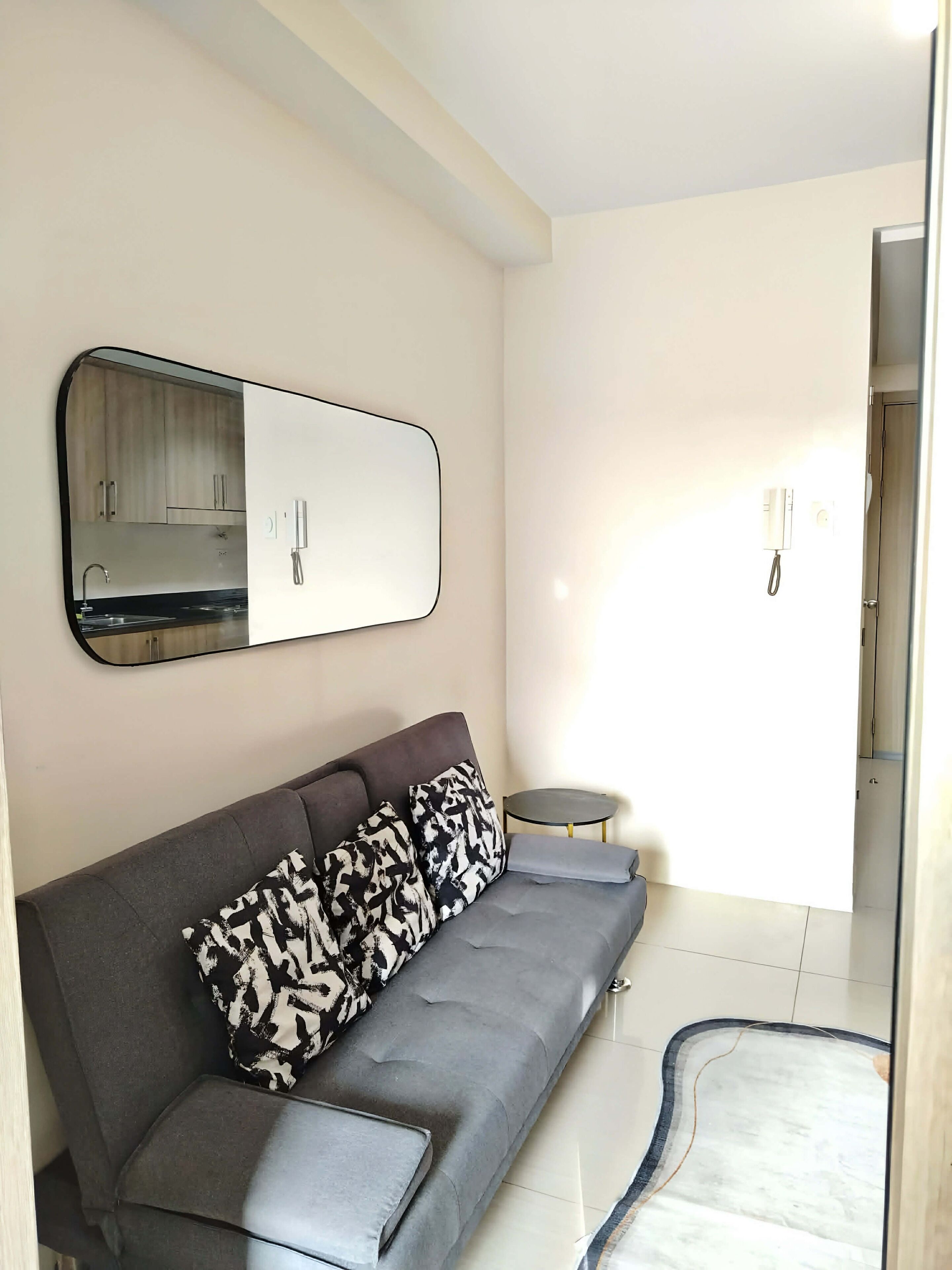 A modern living room with a grey sofa, patterned cushions, a round side table, a large mirror, and a small rug.