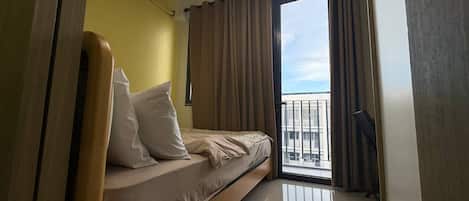 A room with a bed, a window with curtains, a ceiling light, and a view of buildings outside.