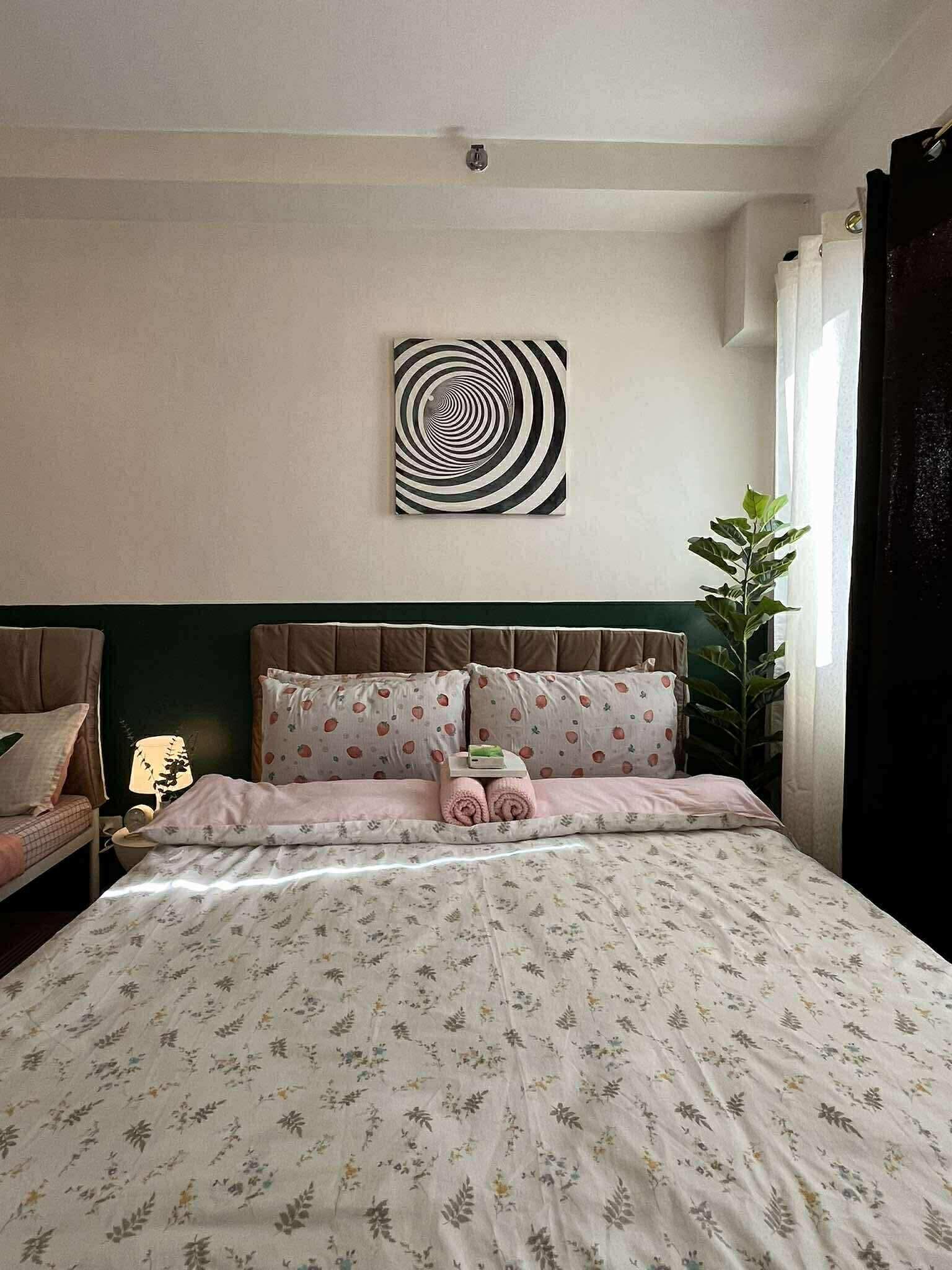 A bedroom with a bed, a headboard, a wall art piece, a nightstand, a lamp, and a potted plant.