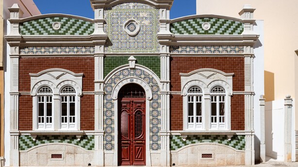 Un bâtiment avec une porte rouge, des encadrements de fenêtres blancs et des carreaux aux motifs verts et bleus.