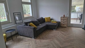 En moderne stue med en grå modulsofa, plankegulv i fiskebensmønster og udsigt til det fri gennem store vinduer.