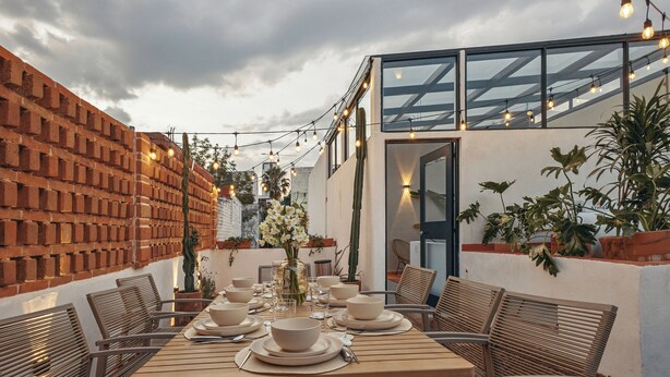 An outdoor dining area with a brick wall, wooden table, and chairs. There are string lights above and potted plants around. - Saltillo Condesa House (Ciudad de México)