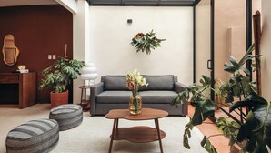 A modern living room with a grey sofa, a wooden coffee table, and two patterned ottomans. There are several potted plants, including a large one on the right and a smaller one on the left. A guitar is leaning against the wall on the left. - Saltillo Condesa House (Ciudad de México)