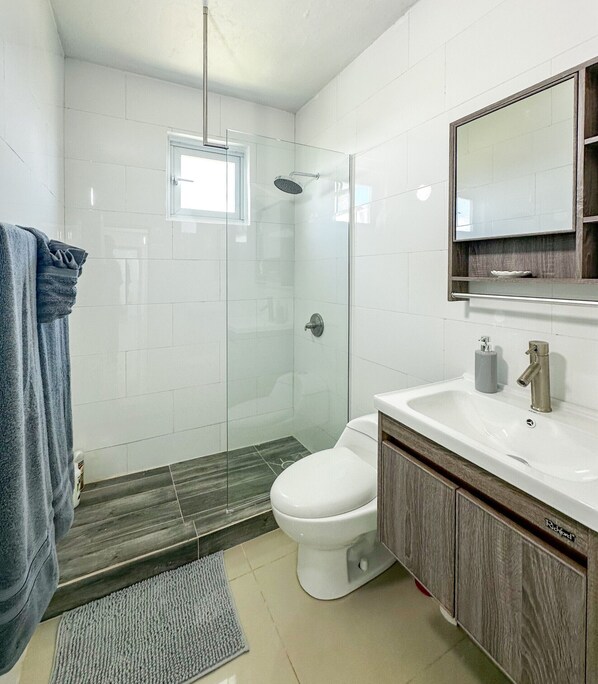 A modern bathroom with a glass-enclosed shower, a white toilet, and a vanity with a mirror. - ARIES APARTMENT (Hatillo)