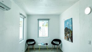 Een kamer met twee zwarte stoelen, een bijzettafeltje met een tas en boeken, een schilderij aan de muur en een raam met uitzicht.