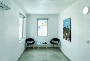A room with two black chairs, a small table with a bag and books, a painting on the wall, and a window with a view of the outside. - ARIES APARTMENT (Hatillo)
