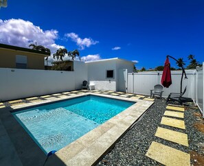 A rectangular swimming pool with a tiled edge, surrounded by a pebble walkway, a white wall, and a red umbrella. - ARIES APARTMENT (Hatillo)