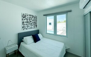 A bedroom with a bed, a nightstand, a wall art piece, and a window with a view of the outside. - ARIES APARTMENT (Hatillo)