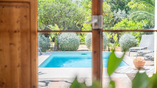 Ein holzgerahmtes Fenster mit Blick auf ein Schwimmbecken, umgeben von üppiger Bepflanzung und einer Terrasse mit Gartenmöbeln.